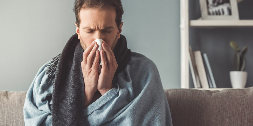 Mann mit Schal und Decke niest in ein Taschentuch auf der Couch, Hintergrund mit Regal und Zimmerpflanze.