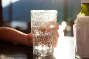 Hand hält Glas mit sprudelndem Wasser, Sonnenlicht im Hintergrund.