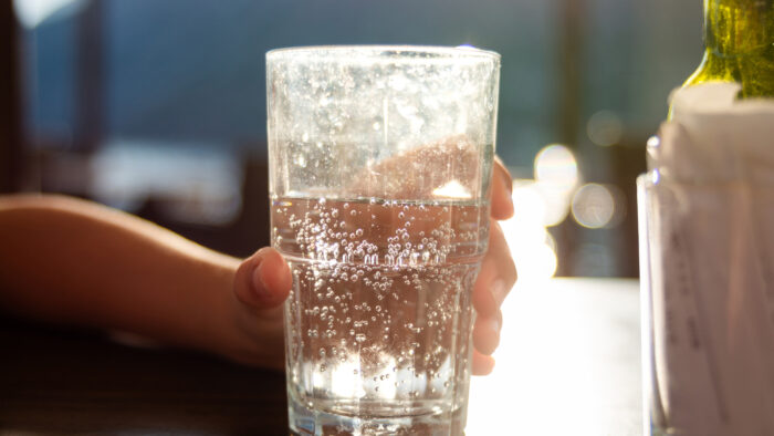 Hand hält Glas mit sprudelndem Wasser, Sonnenlicht im Hintergrund.